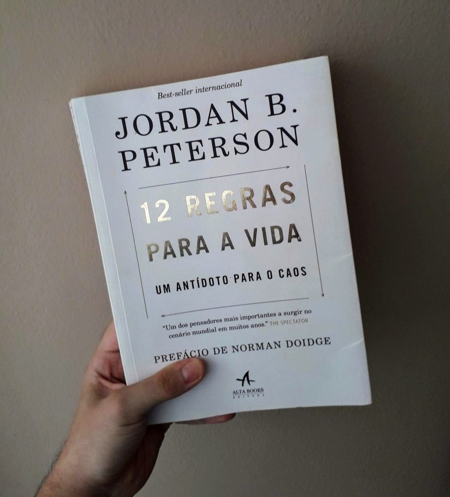 Mão segurando o livro "12 Regras para a Vida: Um Antídoto para o Caos" de Jordan B. Peterson, com capa branca e letras douradas em destaque.
