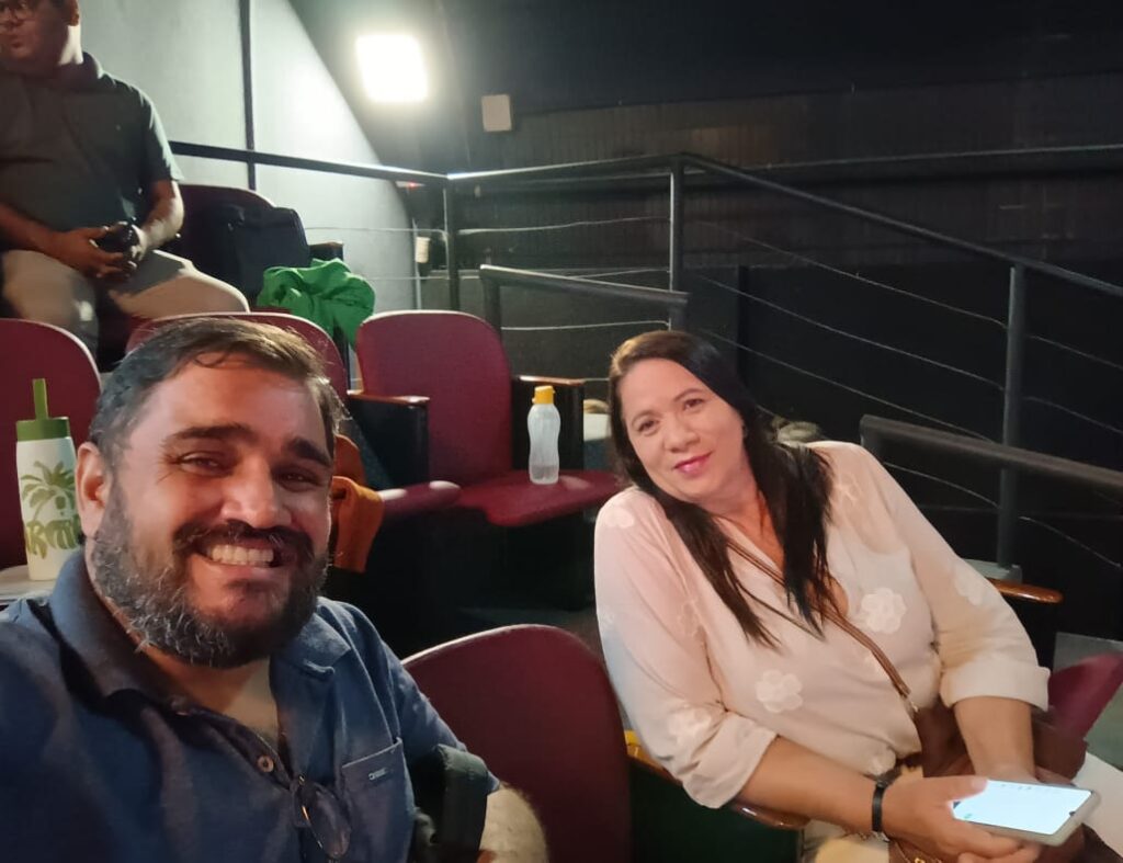 Dois participantes sorridentes fotografados em seus assentos durante a 5ª Mostra Estadual de Práticas de Saúde, compartilhando momento de conexão no auditório.