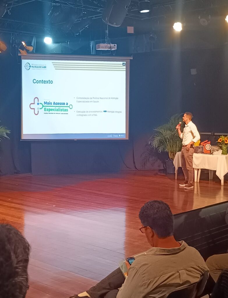 Apresentador explicando o programa "Mais Acesso a Especialistas" durante a 5ª Mostra Estadual de Práticas de Saúde, com slide projetado mostrando contexto da política de atenção especializada.
