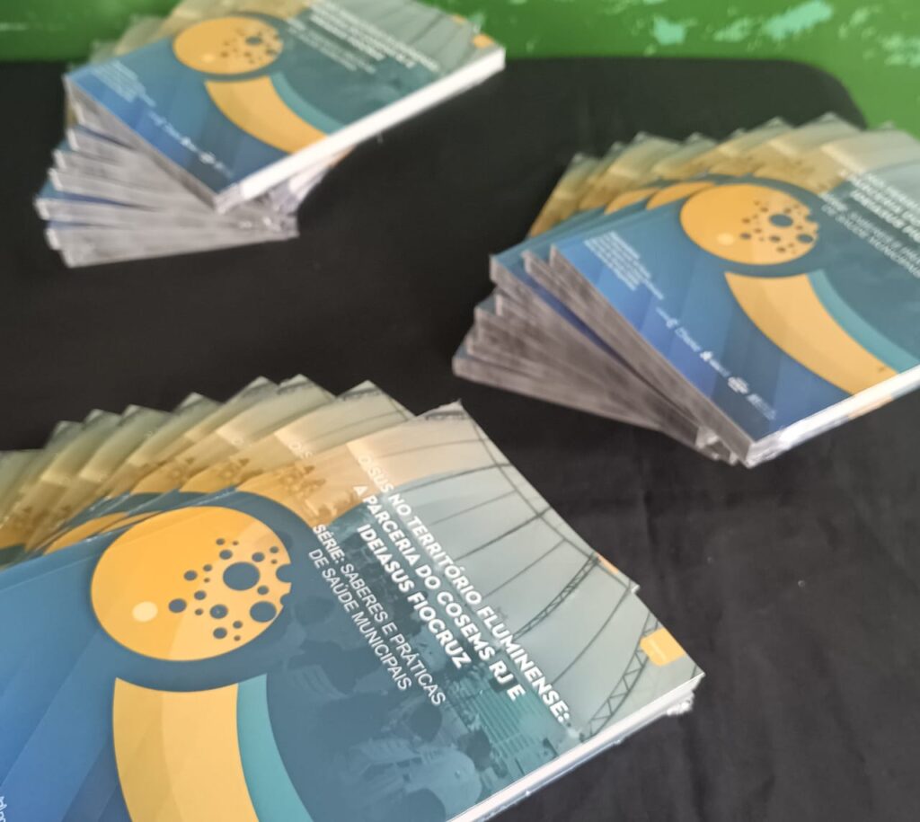 Três pilhas de publicações da série "Saberes e Práticas de Saúde Municipais" do IdeiaSUS/FIOCRUZ e COSEMS RJ sobre uma superfície escura.