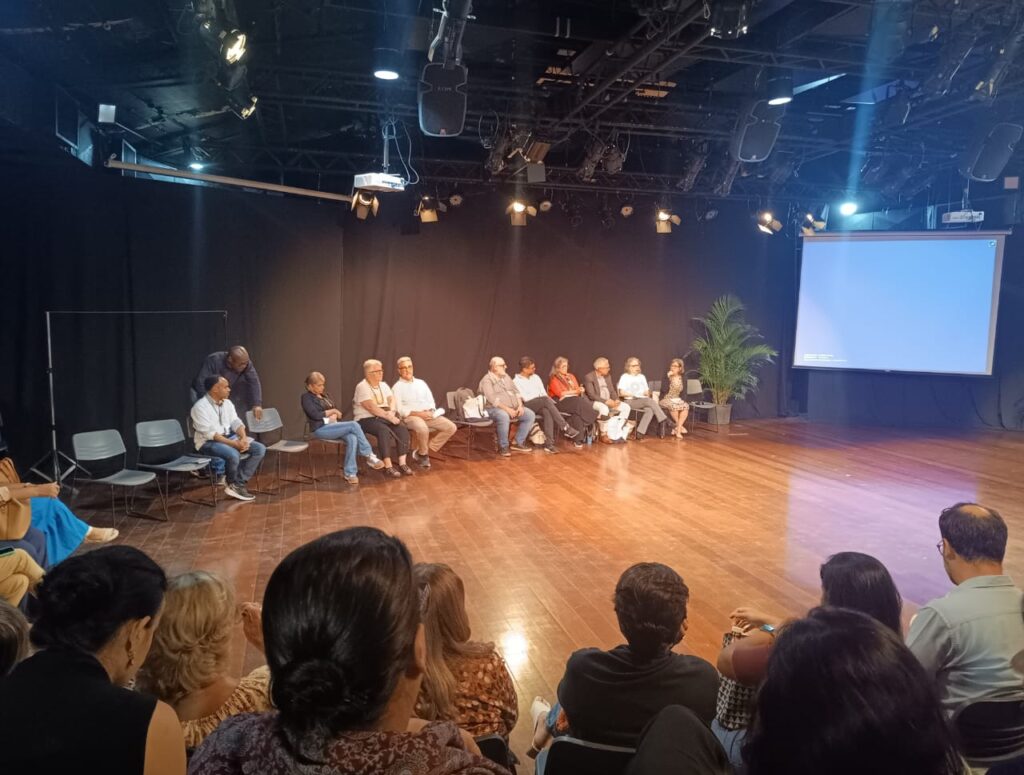 Painel de discussão da 5ª Mostra Estadual de Práticas de Saúde com especialistas sentados em semicírculo no palco e público assistindo em primeiro plano.